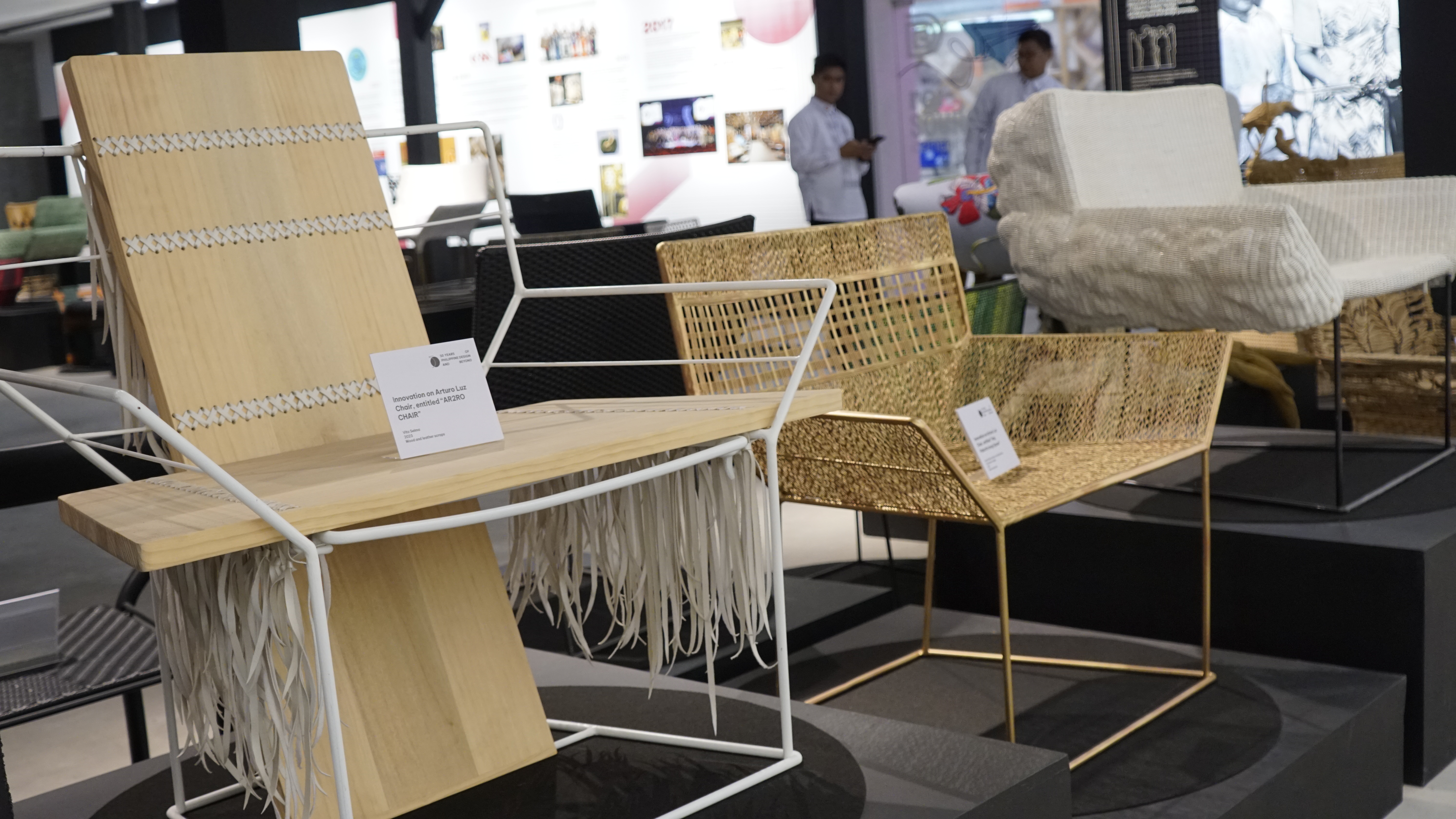A close-up of a modern, light-colored wooden chair with white metal framing and fringe details, displayed on a black platform alongside other woven furniture pieces.