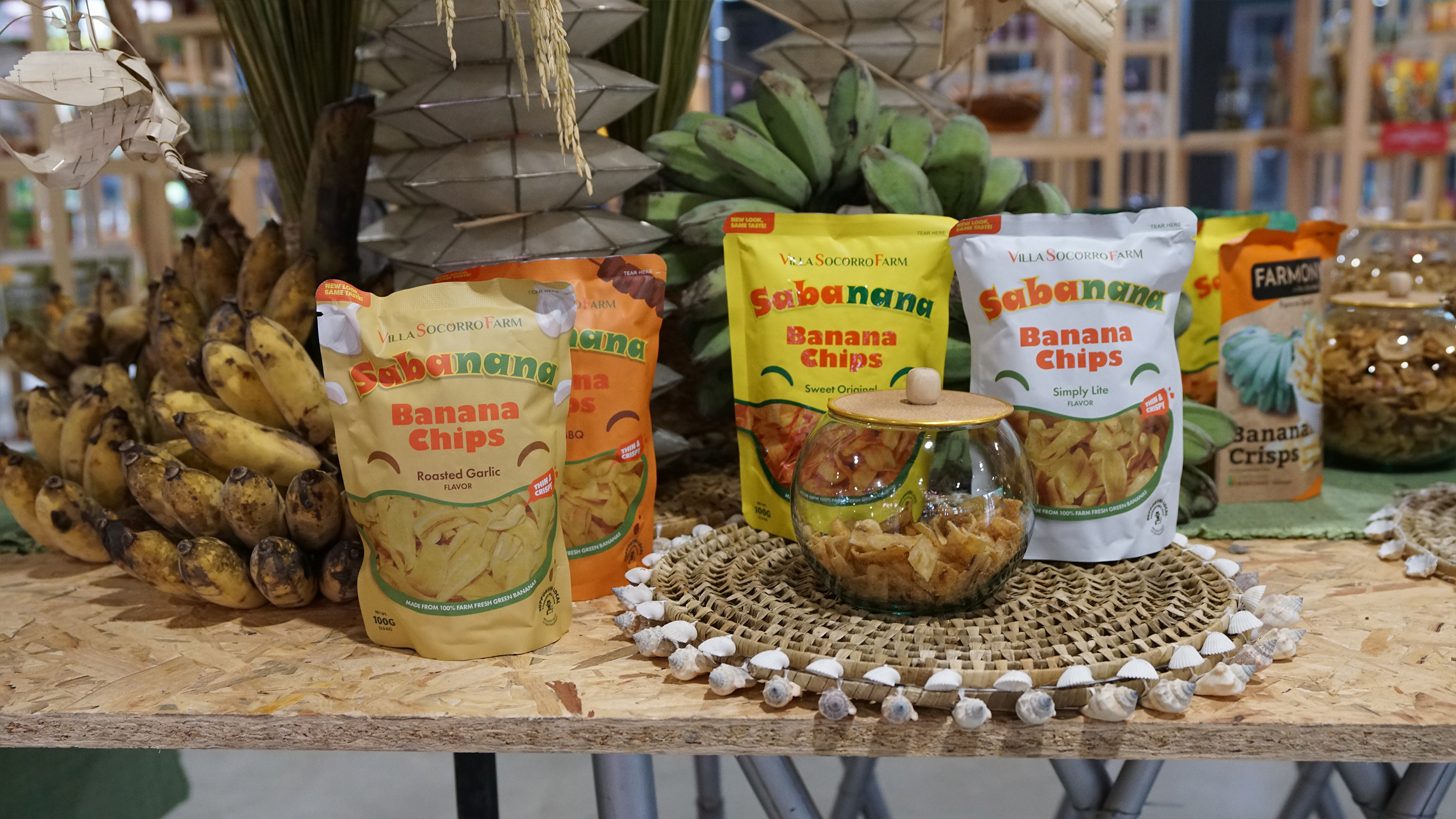 Sabanana banana chip pouches and a glass jar of chips displayed next to fresh green bananas.